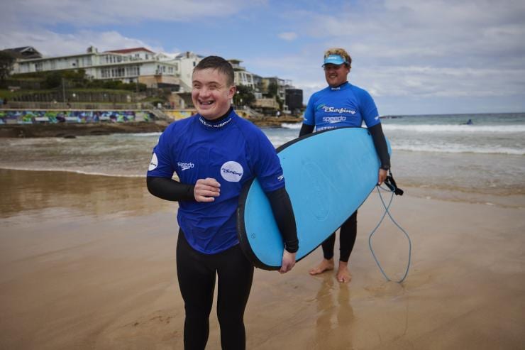 A person holding a blue surfboard walking along the sand with an instructor at Let's Go Surfing, Bondi Beach, New South Wales © Tourism Australia