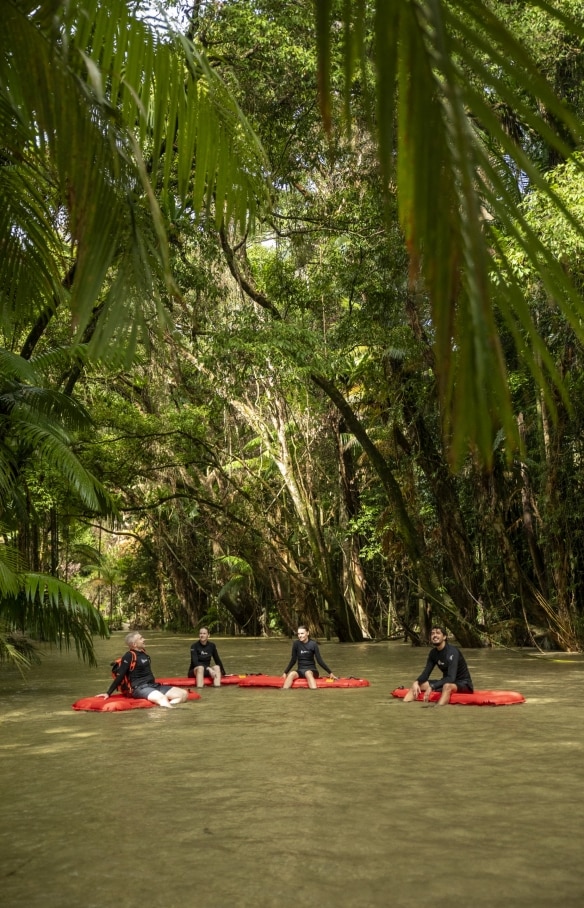 A group of people sitting on inflatable red boards, floating on a calm river beneath a lush green rainforest canopy with Back Country Bliss Adventures, Daintree Rainforest, Queensland © Tourism and Events Queensland