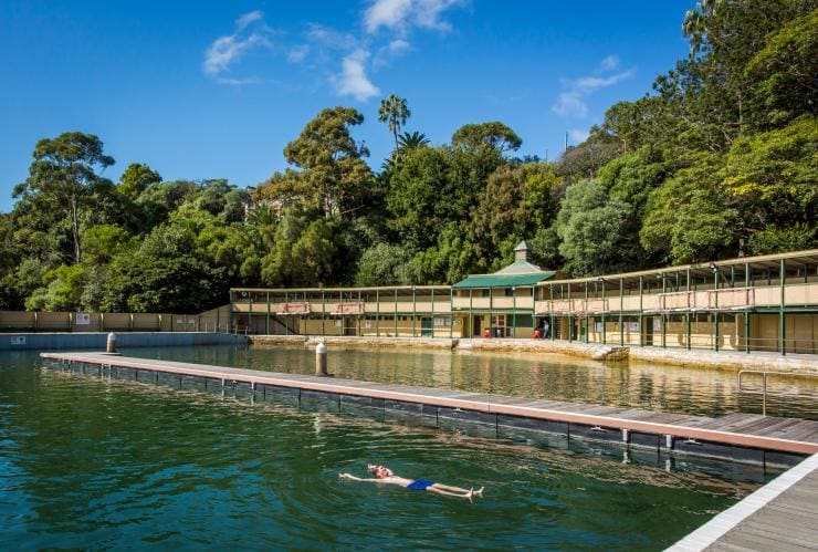 Eine Person schwimmt im blaugrünen Wasser der Dawn Fraser Baths mit den historischen Mauern des von Bäumen umgebenen Gebäudes im Hintergrund, Balmain, Sydney, New South Wales © Destination NSW