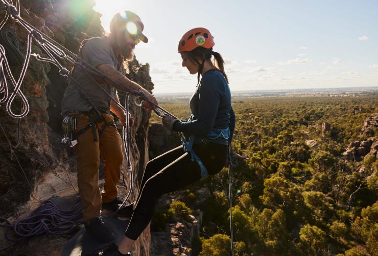 A person abseiling down a sheer cliff overlooking bushland with the help of an instructor with Hangin' Out, Grampians, Victoria © Tourism Australia 