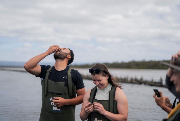 Two people dining on oysters while wearing waders in the ocean at Freycinet National Park, Marine Farm, Freycinet, Tasmania © Tourism Australia