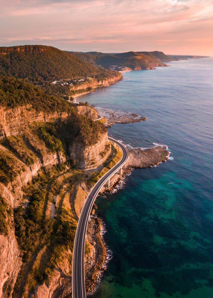 Aerial overlooking cars driving along a bridge jutting out over the ocean on the Sea Cliff Bridge, Clifton, New South Wales © Destination NSW 