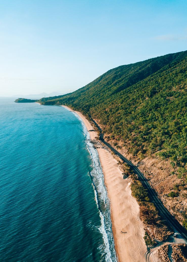 Aerial view over a road running alongside a gold sand beach with a tall mountain covered in rainforest on the other side along the Great Barrier Reef Drive, Tropical North Queensland, Queensland © Tourism and Events Queensland