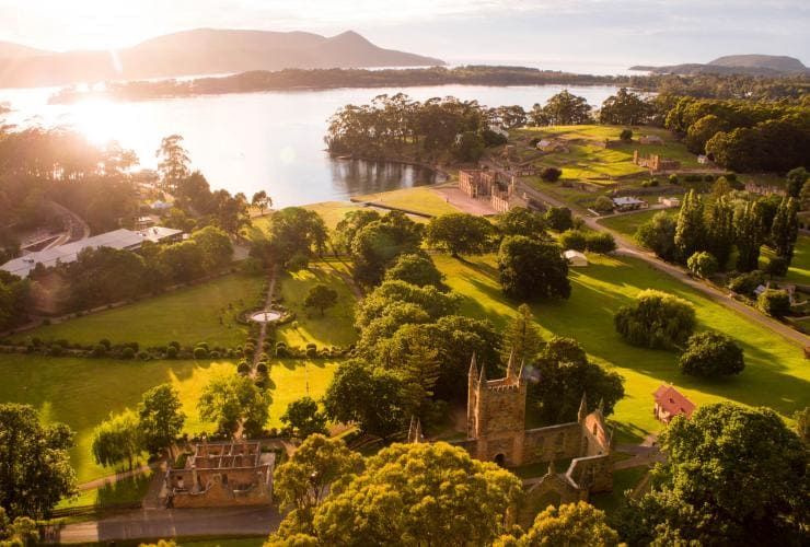 Vue aérienne du soleil éblouissant le site historique de Port Arthur entouré de verdure et d'une eau bleue paisible à Port Arthur, Tasmanie © Alastair Bett