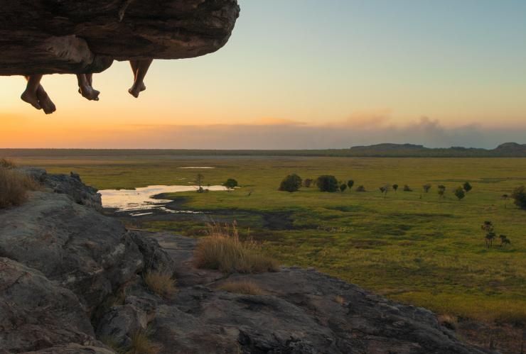 Vue sur les jambes pendant dans le vide de personnes assises sur le rebord d'un site d'art rupestre aborigène à Ubirr, surplombant les étendues de verdure luxuriantes du Kakadu National Park lors du coucher du soleil, Territoire du Nord © Joe Florian