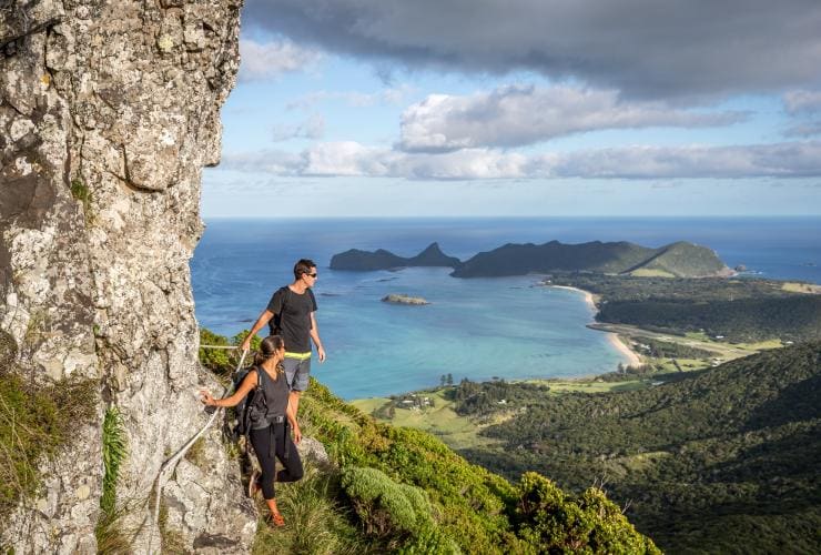Un homme et une femme marchant au bord d'une montagne tout en s'accrochant à une corde, surplombant Lord Howe Island sur le Seven Peaks Walk, Nouvelle-Galles du Sud © GWOA