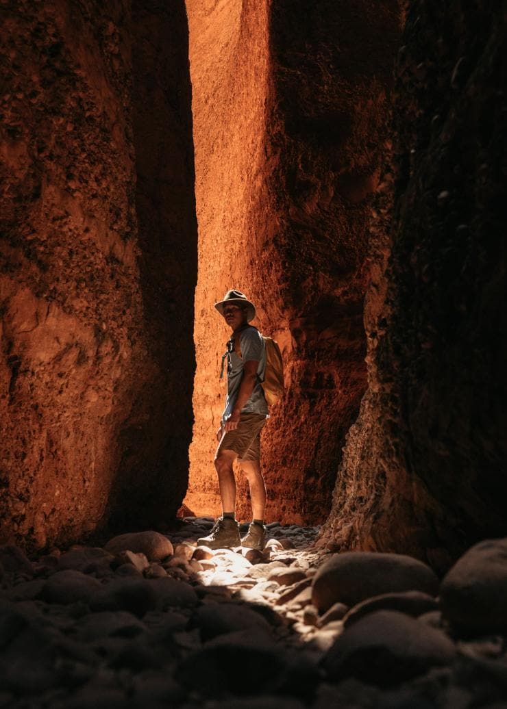 Une personne debout à l'ombre de deux façades rocheuses rouges, avec un faible rayon de soleil derrière elle, à Echidna Chasm, Purnululu National Park, Australie Occidentale © Tourism Western Australia