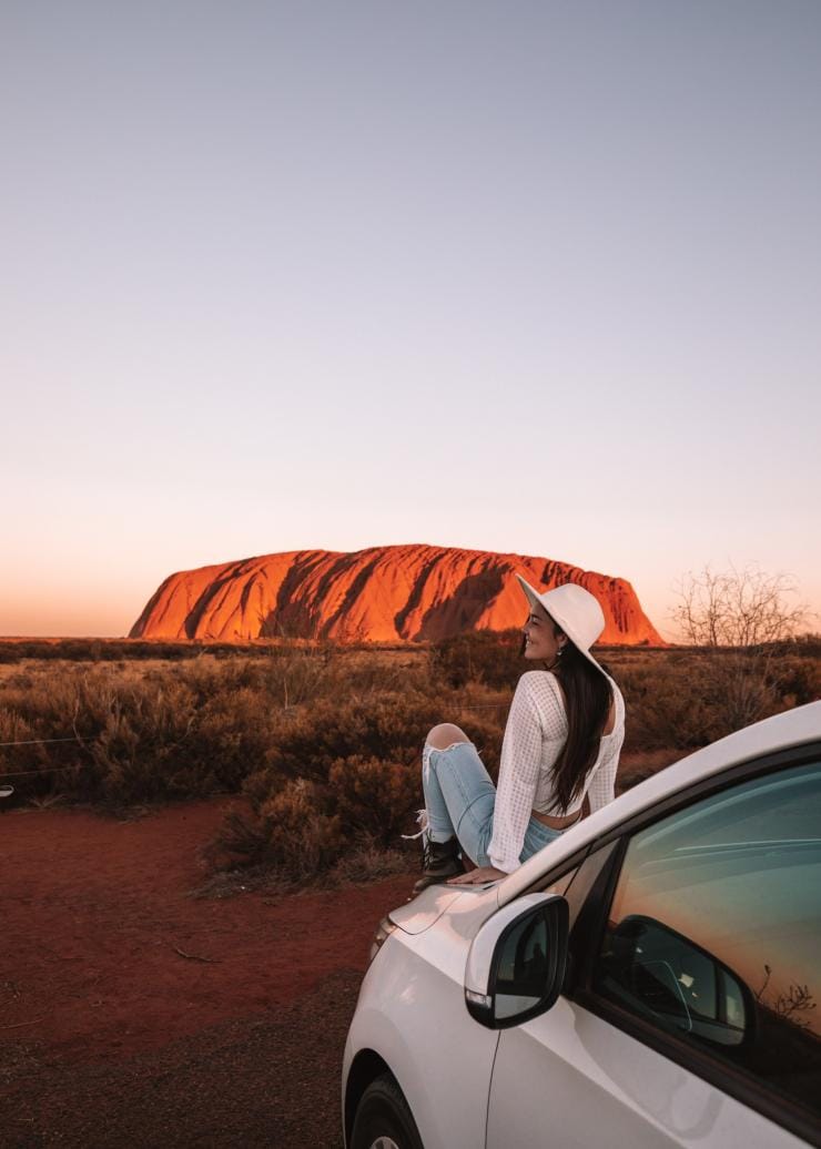 Une femme assise sur le capot d'une voiture, admirant Uluru qui arbore un rouge éclatant lors du coucher du soleil, Uluru-Kata Tjuta National Park, Territoire du Nord © Tourism NT/Lola Hubner