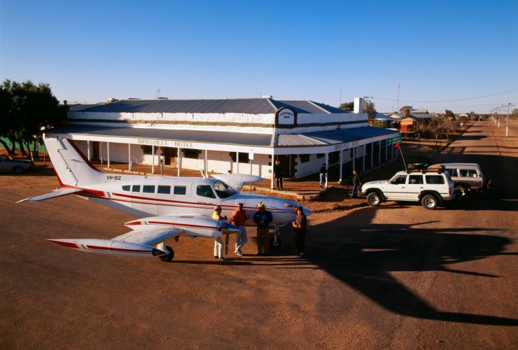 Pemandangan dari udara sebuah pesawat kecil yang terparkir di luar pub bersejarah dengan sekelompok pengunjung sedang berkumpul menikmati minuman dan mengobrol di Birdsville Hotel, Queensland © Tourism Australia