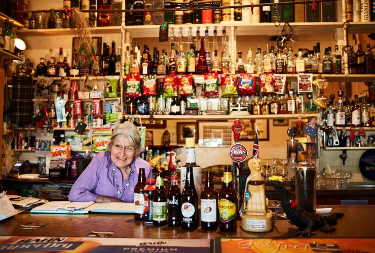 Seorang bartender sepuh tersenyum di bar dengan dikelilingi oleh berbagai botol alkohol di Grand Hotel Kookynie, Kookynie, Western Australia © Tourism Western Australia