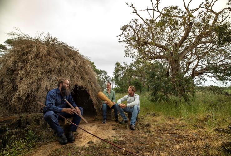 Dua pengunjung bersama pemandu lokal Aborigin sedang duduk di dekat struktur seperti rumah yang terbuat dari cabang-cabang pohon sambil memegang artefak saat tur berpemandu di Budj Bim Cultural Landscape, Victoria © Visit Victoria