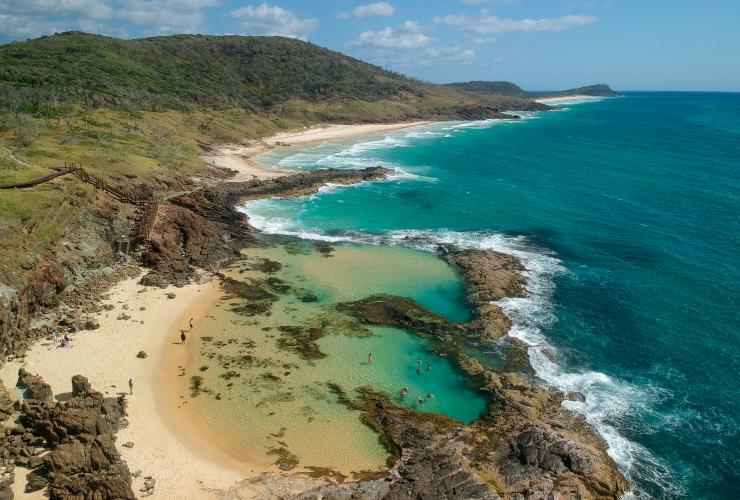Pemandangan dari udara orang-orang yang sedang berenang di kolam formasi karang alami Champagne Pools dengan ombak samudra yang menyentuh tepian kolam di K’gari, Queensland © Tourism Australia
