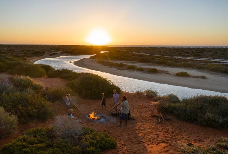 Sebuah keluarga sedang berdiri di sekeliling api unggun bersama pemandu Aborigin lokal di daratan berpasir merah di antara semak rerumputan di sebelah jalur air menjelang matahari terbenam bersama Wula Gura Nyinda Eco Adventures, Shark Bay World Heritage Area, Western Australia © Tourism Western Australia 