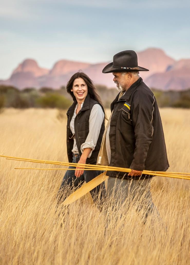 Seorang wanita berjalan melintasi dataran berumput bersama pemandu lokal Aborigin yang memegang artefak saat tur budaya bersama SEIT Outback Australia, Uluru, Northern Territory © Tourism NT/Archie Sartracom
