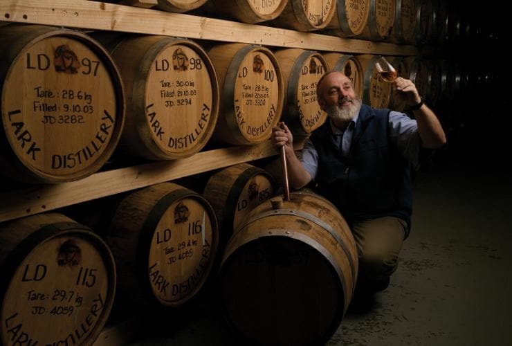 Uomo accovacciato accanto a una parete di botti di rovere che ammira un bicchiere di liquore appena versato alla Lark Distillery, Hobart, Tasmania © Osborne Images