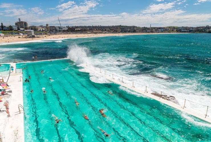 Vista aerea di persone che nuotano nelle acque turchesi della piscina di Bondi Icebergs mentre le onde si infrangono contro i bordi, con Bondi Beach sullo sfondo a Sydney, New South Wales © Destination NSW