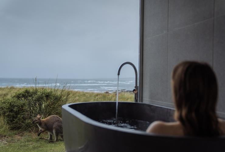 Una persona seduta in una grande vasca da bagno davanti a una vetrata a tutta altezza da cui è possibile osservare un wallaby e l'oceano in lontananza al Kittawa Lodge, King Island, Tasmania © Emilie Ristevski